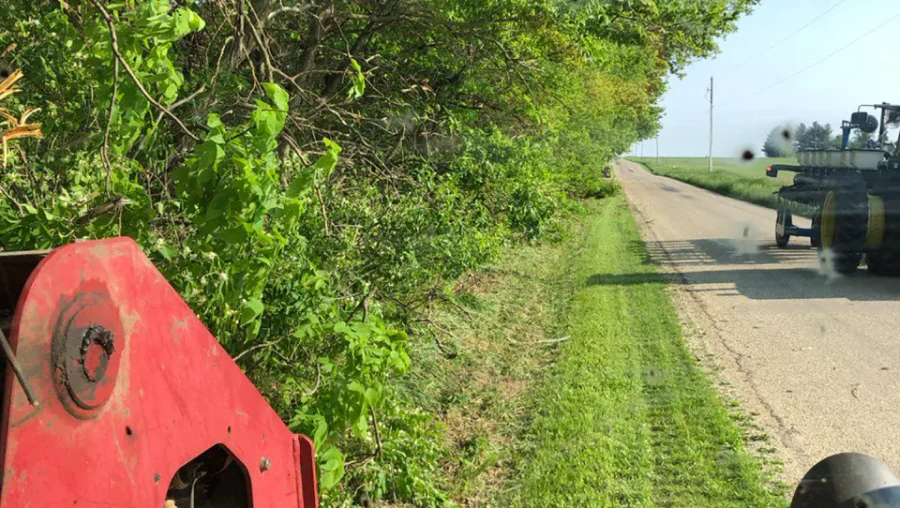 A heavy-duty red tractor with a cutting attachment clearing overgrown grass and bushes beside a rural road